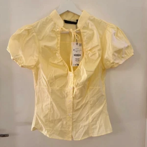 Gul puffärmad blus med knyt från zara - Söt ljusgul blus med puffiga korta ärmar och knytband vid halsen. Blusen har knäppning framtill och en feminin, figurnära passform. Tillverkad i ett lätt bomullsliknande material som känns fräscht och somrigt.