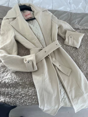 Enzaro elegant lång cremefärgad kappa med skärp - Stilren beige trenchcoat från Enzaro med klassisk krage, knytskärp i midjan och dekorativa detaljer vid ärmsluten. Jackan har en mjuk, mockaliknande yta och quiltat innerfoder för extra komfort. Perfekt för dig som vill ha en tidlös och trendig look. Passar Storlek S/M