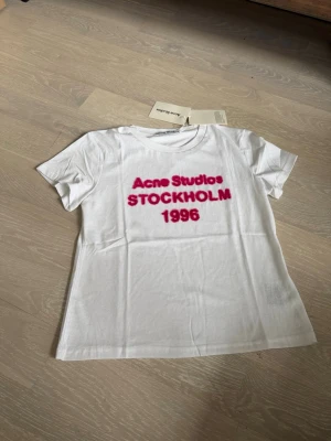 Vit Acne Studios t-shirt med tryck - Vit t-shirt från Acne Studios med rosa text 'Acne Studios STOCKHOLM 1996' på bröstet. Klassisk rund halsringning och korta ärmar. Perfekt för dig som gillar stilrena plagg med en twist.