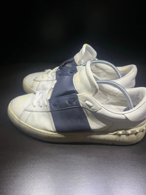 Vita och marinblå sneakers Valentino - Snygga sneakers från Valentino i vitt läder med en bred marinblå rem över sidan. Klassisk låg modell med vita snören och detaljerad sula. Perfekta för dig som vill ha en stilren men ändå unik look.