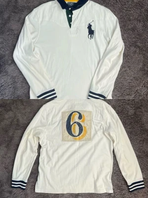 Vit långärmad rugby tröja från Polo Ralph Lauren - Vit långärmad rugby tröja från Polo Ralph Lauren med mörkblå krage och knappar. Stort mörkblått logotryck på bröstet och en stor gul-blå siffra 6 på ryggen. Randiga muddar vid ärmsluten ger en sportig touch.