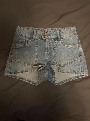 Ljusblå denimshorts med broderade fickor - Low waist ljusblå denimshorts och snygga broderade hjärtan på bakfickorna. Shortsen har framfickor, bälteshällor och dragkedja med knapp. Använt 1 gång.