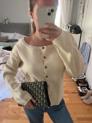 Krämvit kofta med knappar PRISLAPP KVAR - Stilren krämvit kofta med rund halsringning och dekorativa knappar framtill. Koftan har lång ärm och en rak, avslappnad passform. Perfekt att matcha med jeans för en enkel men snygg look.