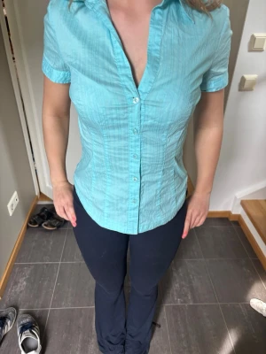 Turkos tight skjorta - Säljer en ljusblå skjorta med korta ärmar och klassisk krage. Skjortan har knappar framtill och är tillverkad i ett lätt, något strukturerat bomullstyg. Perfekt för en fräsch och enkel stil.