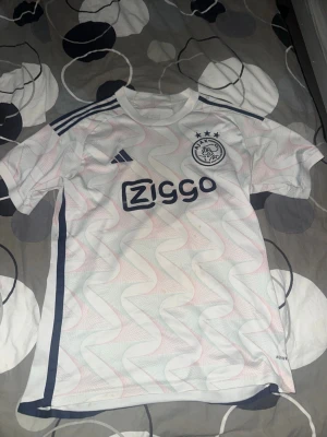 Ajax vit matchtröja Adidas - Säljer en vit Ajax matchtröja från Adidas med marinblå detaljer och Ziggo-logga på bröstet. Tröjan har ett vågigt mönster i ljusrosa och ljusblått, klubbmärke på vänster bröst och tre klassiska Adidas-ränder på axlarna. Tillverkad i lätt och ventilerande material.