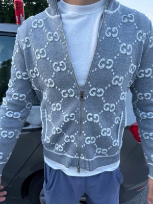 Hm tröja  - Snygg grå ziptröja med vitt GG-mönster över hela plagget. Tröjan har hög krage, ribbade muddar och dragkedja framtill. Perfekt för dig som gillar stilrena detaljer och vill sticka ut lite extra.