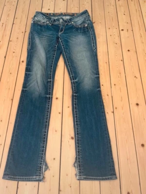 Rock Revival bootcut jeans blå - Säljer ett par blå bootcut jeans från Rock Revival med snygga slitningar och kontrastsömmar. Bakfickorna har broderade detaljer och silverknappar. Jeansen har låg midja och klassisk femficksmodell. Perfekta för dig som gillar en avslappnad men trendig stil.