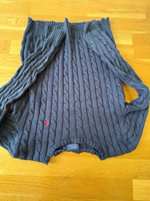 Mörkblå kabelstickad tröja Polo - Mörkblå kabelstickad tröja från Polo Ralph Lauren med rund halsringning och lång ärm. Tröjan har ett klassiskt kabelmönster och en liten röd broderad logga på bröstet. Perfekt för en stilren och avslappnad look.