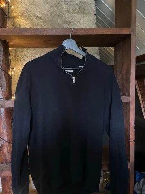 Svart halvzip tröja Jack & Jones - Svart stickad tröja från Jack & Jones med halvzip och hög krage. Perfekt för lager på lager och enkel att matcha med olika outfits. Tillverkad i mjukt material och har långa ärmar samt ribbade muddar.