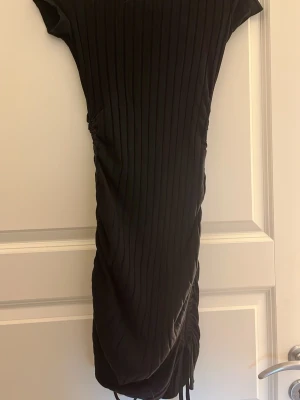 Svart ribbad kortklänning - Snygg svart kortklänning med ribbad struktur och snörning i sidan. Klänningen har korta ärmar och en kroppsnära passform som framhäver figuren. Perfekt för dig som gillar stilrena och trendiga plagg.