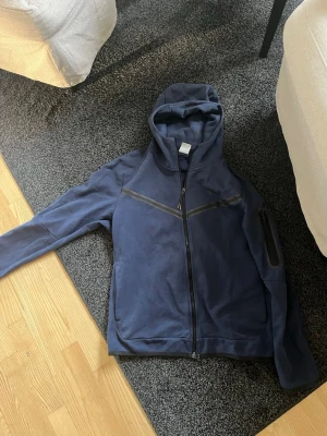 Nike tech  - Snygg mörkblå hoodie med hel dragkedja framtill och huva. Hoodien har en diagonal detaljsöm över bröstet och en ficka med dragkedja på ena ärmen. Perfekt för en avslappnad och sportig stil. Obs! Det är en Nike tech så fråga ej. Bra skick.