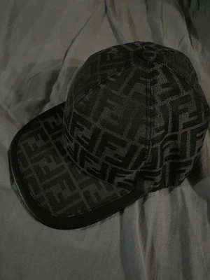 Fendi keps - Snygg svart keps från Fendi med det klassiska FF-logomönstret över hela kepsen. Kepsen har böjd skärm med läderdetalj och justerbart band bak. Perfekt accessoar för dig som gillar exklusiv streetwear och vill sticka ut.