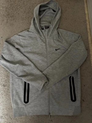 Grå zip hoodie från Nike - Snygg grå hoodie från Nike med dragkedja framtill och svart Nike-logga på bröstet. Hoodien har två fickor med svarta detaljer och en klassisk huva. Perfekt för en sportig och avslappnad stil.