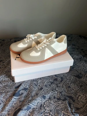 Vita och beige sneakers från Arveilo - Stilrena sneakers i vitt och beige från Arveilo med klassisk retrodesign. Ovandelen är i en mix av mocka och slätt material, med vita snören och en brun gummisula som ger skorna en snygg kontrast. Perfekta för dig som gillar minimalistisk och tidlös stil. Oanvända