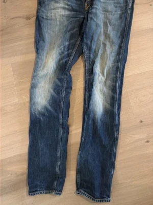 Blå raka jeans med slitningar - Säljer ett par blå jeans med raka ben och tydliga slitningar på framsidan. Jeansen har klassisk femficksdesign och kontrastsömmar. Materialet är denim i bomull och färgen är tvättad blå med ljusare partier på lår och knän. Längd 32 bredd 34. Kan gå ner till 300 ungefär vid snabb affär. 