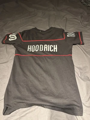 Svart HOODRICH t-shirt med röda detaljer - Svart t-shirt från HOODRICH med vita tryck på bröstet och ärmarna. T-shirten har röda sömmar som ger en schysst kontrast och en modern streetwear-look. Klassisk passform och rund hals. Perfekt för dig som gillar stilrena men ändå unika plagg.
