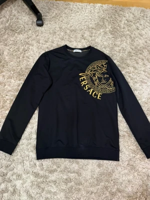 Svart sweatshirt från Versace - Svart långärmad sweatshirt från Versace med stort guldigt Medusa-tryck och logga på bröstet. Rund halsringning och ribbade muddar vid ärmslut och nederkant. Versace-logga även på ena ärmen. Perfekt för dig som gillar exklusiv streetwear.