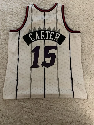 Toronto Raptors Vince Carter tröja - Säljer en vit basketlinne från Toronto Raptors med Vince Carter och nummer 15. Tröjan har svarta och röda detaljer, blixtmönster och stor röd dinosaurie på framsidan. Tillverkad i meshmaterial som andas, perfekt för basket eller streetwear.
