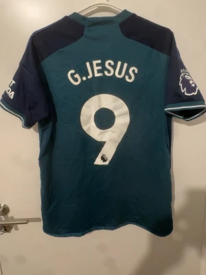 Arsenal bortatröja G.Jesus #9 - Snygg Arsenal bortatröja från Adidas med G.Jesus och nummer 9 på ryggen. Tröjan är mörkgrön med mörkblå ärmar och vita detaljer. Premier League-logga på ärmen och Emirates Fly Better-tryck på bröstet. Perfekt för match eller träning.