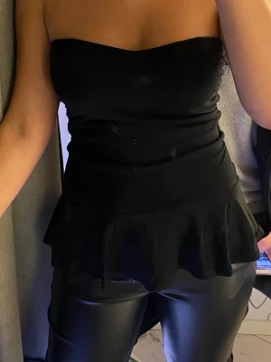 Svart axelbandslös peplum tubtopp - Säljer en svart axelbandslös tubtopp med peplumvolang nedtill. Toppen har en tight passform och är perfekt för dig som vill ha en stilren och trendig look. Materialet känns mjukt och följsamt mot kroppen.