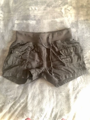 Svarta Cargoshorts H&M – Retro/Y2K strl 34 - Svarta cargoshorts från H&M i 100% bomull med stretchig trikåmudd och knytsnöre i midjan. Klockren retro/Y2K-stil! Totalt 6 funktionella fickor (4 fram, 2 bak m. knapp). Mått: • Midja platt: 34,5 cm (stretchig mudd) • Innerben (från skrevet): 8 cm • Totallängd (midja till benslut): 31 cm Storlek 34 (XS). Använt skick!