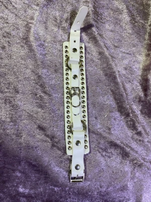 Vitt nitar armband  - Säljer ett vitt armband i skinnimitation med silverfärgade nitar och ringar. Bältet har en bred design och justerbart spänne. Perfekt för att ge din outfit en edgy touch.helt ny 