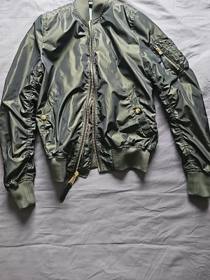 Olivgrön bomberjacka Alpha Industries - Snygg olivgrön bomberjacka från Alpha Industries med klassisk glansig finish, ribbade muddar och dragkedja framtill. Jackan har fickor med tryckknappar och en ärmficka med dragkedja. Perfekt för dig som vill ha en tidlös och cool look. Nypris 2495
