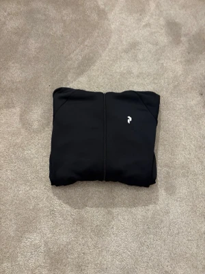 Svart hoodie med dragkedja och tryck - Säljer en svart peak performance hoodie med dragkedja och huva. På bröstet finns en liten vit logga och i huvan står det 'PEAKPERFORMANCE' i vitt. Hoodien har två fickor med dragkedja och är gjord i ett mjukt material. Perfekt för en chill och stilren look.