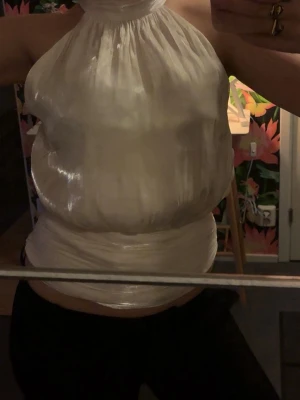 Vit halterneck topp i glansigt material - Säljer en unik vit halterneck topp med glansig finish och hög hals. Toppen är ärmlös och har en lös passform med inslag av omlott runt midjan. Perfekt för dig som vill sticka ut med en cool och trendig look.