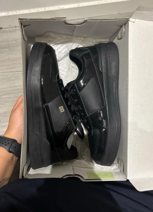 Tommy hilfiger skor - Snygga svarta sneakers med blank finish och låga kanter. Skorna har svarta snören och en diskret gulddetalj på sidan. Tillverkade i en mix av syntetmaterial och läderimitation för en clean look. Perfekta för dig som gillar stilrena och moderna skor. Helt oanvända!