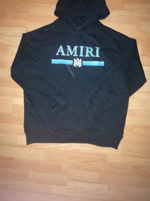 Svart hoodie med AMIRI - Snygg svart hoodie från AMIRI med stort vitt tryck på bröstet och blåa linjer under loggan. Klassisk huva och känguruficka framtill. Perfekt för dig som gillar streetwear och vill ha en stilren men ändå iögonfallande look.