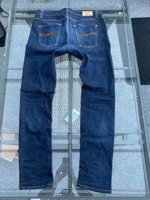Nudie Jeans, Lean Dean - Dry 16 Dips 33/34 - Säljer ett par mörkblå jeans från Nudie Jeans med klassisk gul söm på bakfickorna och patch med logga i midjan. Jeansen har en slim passform och är tillverkade i bomull. Perfekta för dig som gillar stilrena och tidlösa jeans.