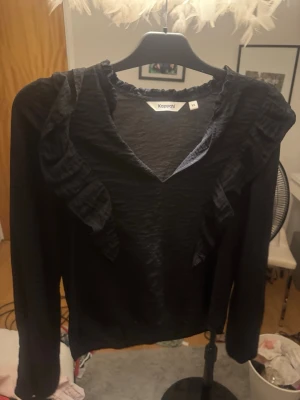 Svart blus med volang från Kappahl - Svart blus från Kappahl med v-ringning och dekorativa volanger längs ringningen och axlarna. Tillverkad i ett tunt, lätt skrynkligt tyg som ger en avslappnad men ändå chic känsla. Långärmad modell som passar perfekt till jeans eller kjol.