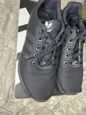 Svarta Adidas sneakers - Snygga svarta Adidas sneakers med klassiska tre ränder på sidan och Torsion-systemet på plösen. Ovandelen är i slitstark mesh och skorna har svarta skosnören. Perfekta för dig som gillar stilrena och sportiga sneakers.