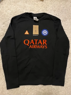 NIKE X Inter Milan Fotball Tröja - Snygg svart långärmad tröja från Nike med Inter-logga och färgglada detaljer. Framsidan har tryck med Qatar Airways och ACG-logga, baksidan har broderad orm och texten 'Football' i orange. Perfekt för dig som gillar fotboll och streetwear.