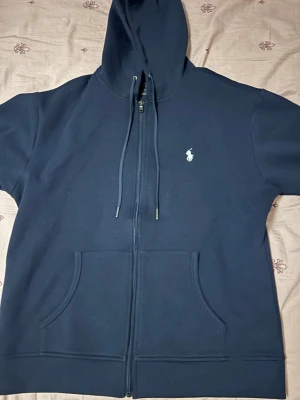 Mörkblå zip hoodie med huva - Storlek L. Snygg mörkblå hoodie med dragkedja och huva. Klassisk design med fickor framtill och justerbara snören i huvan. Liten vit logga broderad på bröstet. Perfekt för en avslappnad stil och enkel att matcha med andra plagg.