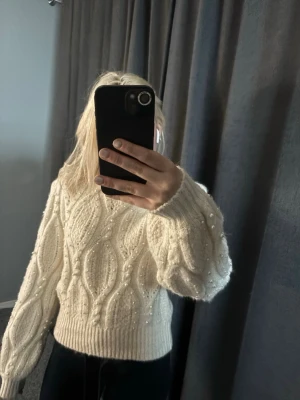 Vit kabelstickad tröja från Zara - Säljer en vit kabelstickad tröja från Zara med pärldetaljer. Tröjan har en croppad passform, ribbade muddar och rund hals. Perfekt för dig som vill ha en mysig och trendig look till hösten eller vintern.