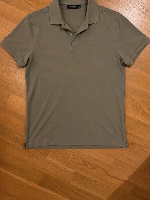 Olivgrön J Lindberg piké - Snygg olivgrön pikétröja med klassisk krage och korta ärmar. Tröjan har två knappar vid halsen och är tillverkad i ett mjukt, stretchigt material som sitter skönt på kroppen. 