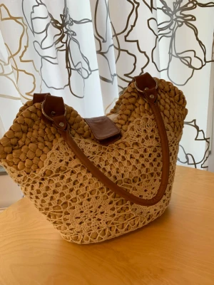 Beige virkad handväska med läderdetaljer - Snygg beige handväska med virkat mönster och bruna läderhandtag. Väskan har en unik struktur med stora virkade bubblor upptill och ett blommigt spetsmönster nedtill. 