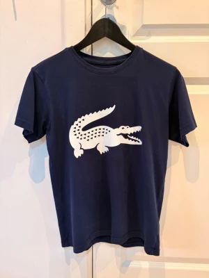 Mörkblå t-shirt med krokodiltryck - Snygg mörkblå t-shirt med stort vitt krokodiltryck på bröstet. Klassisk rund hals och korta ärmar. Perfekt för dig som gillar stilrena plagg med en twist. Materialet känns mjukt och skönt mot huden.