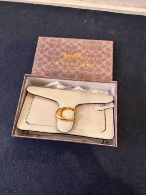 Vit kuvertväska från Coach - Snygg vit kuvertväska från Coach i strukturerat läder med guldfärgat C-spänne framtill. Väskan har svarta kanter och klassisk, stilren design. Levereras i originalbox och med dustbag från Coach.