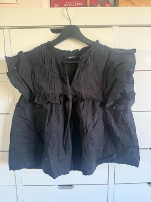Svart blus med volanger från Gina Tricot - Svart blus från Gina Tricot med korta volangärmar och rynkade detaljer. Blusen har rundad halsringning med knytband och dekorativa veck framtill. Tillverkad i ett lätt tyg som ger en luftig känsla. Perfekt för en trendig och avslappnad look.