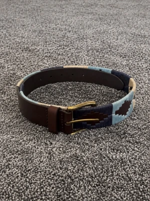 Brunt bälte med blått mönster - Snyggt bälte i brunt skinn med detaljer i blått, beige och mörkblått mönster. Bältet har en klassisk metallspänne i guldton och passar perfekt för att lyfta din outfit med en unik touch.