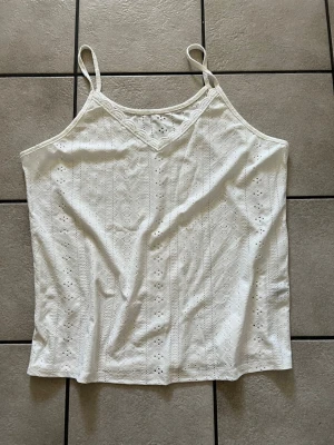 Vit linnetopp storlek XL - Söt vit linnetopp med tunna axelband och v-ringning. Toppen har broderade detaljer och spetsmönster som ger en romantisk känsla. Perfekt för varma dagar och enkel att matcha med jeans eller kjol.
