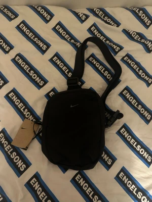 Svart axelväska från Nike - Snygg svart axelväska från Nike med justerbar rem och minimalistisk design. Väskan har ett litet Nike-logga framtill och praktiska fack för dina viktigaste grejer. Perfekt för att bära med sig mobilen, plånboken och andra småsaker.