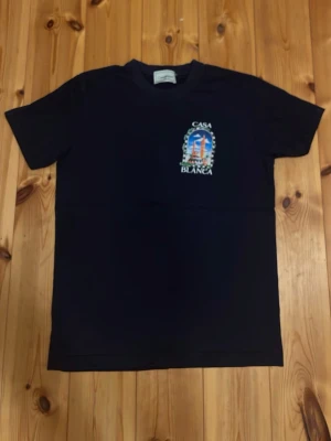 Mörkblå t-shirt från Casablanca - Mörkblå t-shirt från Casablanca med färgglatt tryck på bröstet där det står 'Casa Blanca' och ett motiv med palmer och byggnader. Klassisk passform och rund halsringning. Perfekt för en chill och stilren look.