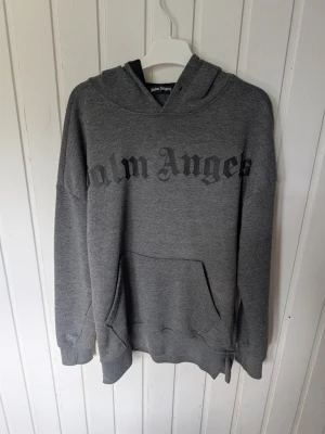 Grå hoodie från Palm Angels - Säljer en grå hoodie från Palm Angels med stor svart logga över bröstet. Hoodien har huva, magficka och ribbade muddar vid ärmar och nederkant. Tillverkad i mjukt bomullsmaterial, perfekt för en avslappnad streetwear-look.