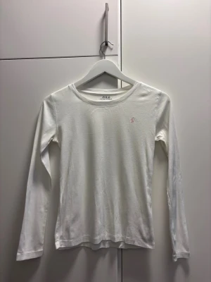 Vit långärmad topp från Polo Ralph Lauren - Klassisk vit långärmad topp från Polo Ralph Lauren med rund halsringning och diskret rosa logga broderad på bröstet. Tillverkad i mjuk bomull som känns skön mot huden. Perfekt att styla med jeans eller kjol för en clean look.