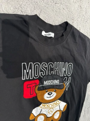 Svart Moschino t-shirt med nalle - Snygg svart t-shirt från Moschino med ett stort broderat motiv av en nallebjörn på framsidan. Nallen har en keps och t-shirt med Moschino-logga. T-shirten har rund hals och korta ärmar, tillverkad i mjuk bomull. Perfekt för dig som gillar streetwear och lekfull design.