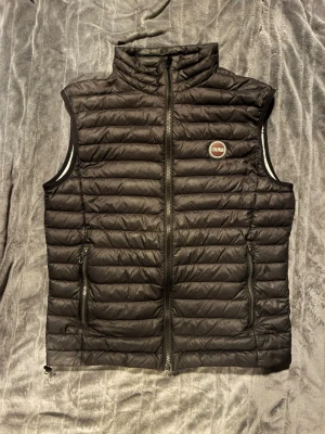 Svart dunväst från Colmar - Snygg svart dunväst från Colmar med quiltad design och hög krage. Västen har två dragkedjeförsedda fickor och en tydlig Colmar-logga på bröstet. Insidan är ljus och västen stängs med dragkedja framtill. Perfekt för lager-på-lager och streetwear.